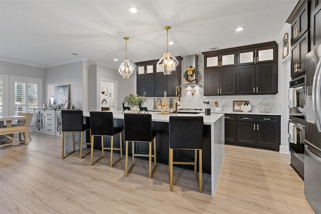 kitchen with tasteful backsplash, wall chimney range hood, light hardwood / wood-style flooring, hanging light fixtures, and an island with sink