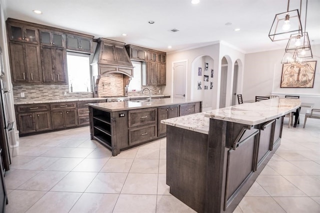 kitchen with dark brown cabinets, a center island with sink, hanging light fixtures, and crown molding