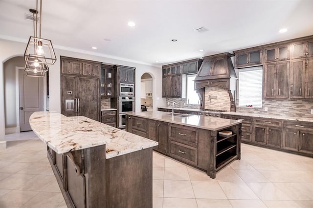 kitchen with custom range hood, built in appliances, hanging light fixtures, and a spacious island