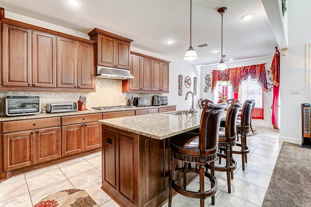 kitchen featuring stainless steel gas cooktop, light stone counters, light tile patterned flooring, hanging light fixtures, and a kitchen island with sink