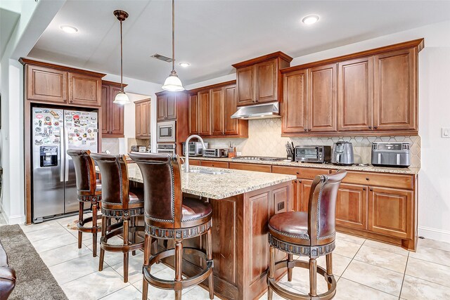 kitchen with a center island with sink, stainless steel appliances, light stone countertops, pendant lighting, and backsplash