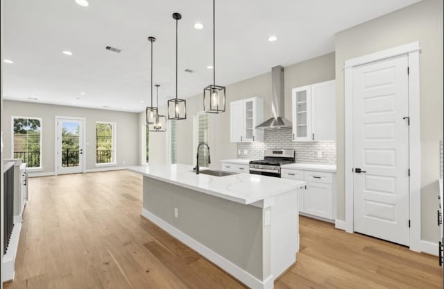 kitchen with sink, wall chimney range hood, stainless steel gas range oven, an island with sink, and white cabinets