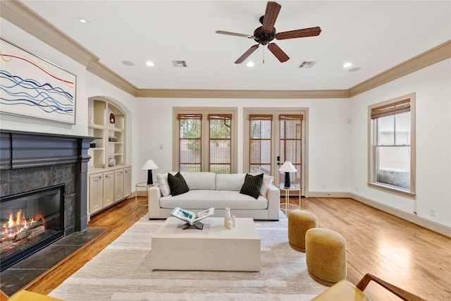 living room with ornamental molding, ceiling fan, a tile fireplace, light hardwood / wood-style flooring, and built in features