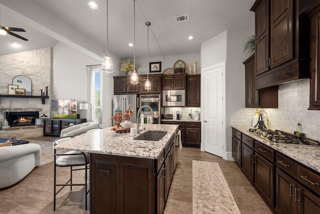 kitchen with a stone fireplace, dark brown cabinets, an island with sink, pendant lighting, and stainless steel appliances