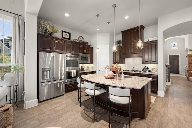 kitchen featuring an island with sink, pendant lighting, stainless steel appliances, light stone countertops, and decorative backsplash