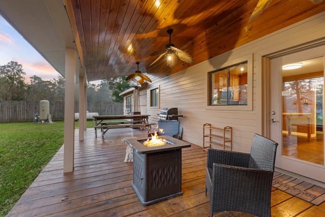 deck at dusk featuring grilling area, a fire pit, ceiling fan, and a lawn