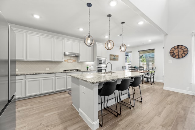 kitchen with light wood-type flooring, light stone counters, a kitchen island with sink, white cabinetry, and hanging light fixtures