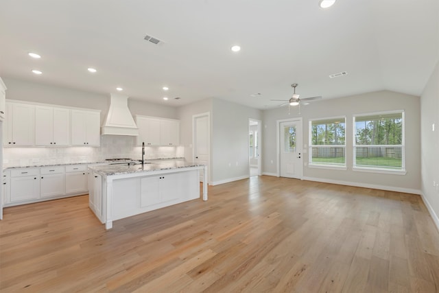 kitchen with white cabinetry, light hardwood / wood-style flooring, backsplash, a center island with sink, and custom exhaust hood