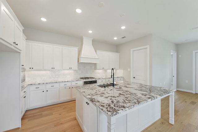 kitchen with custom exhaust hood, light hardwood / wood-style flooring, sink, and an island with sink