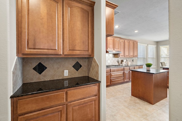 kitchen featuring stainless steel gas stovetop, a center island, backsplash, dark stone counters, and light tile patterned floors