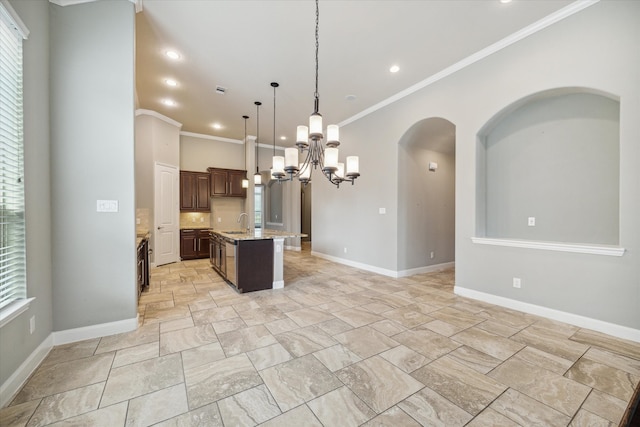 kitchen with light stone countertops, dark brown cabinetry, a kitchen island with sink, crown molding, and decorative light fixtures