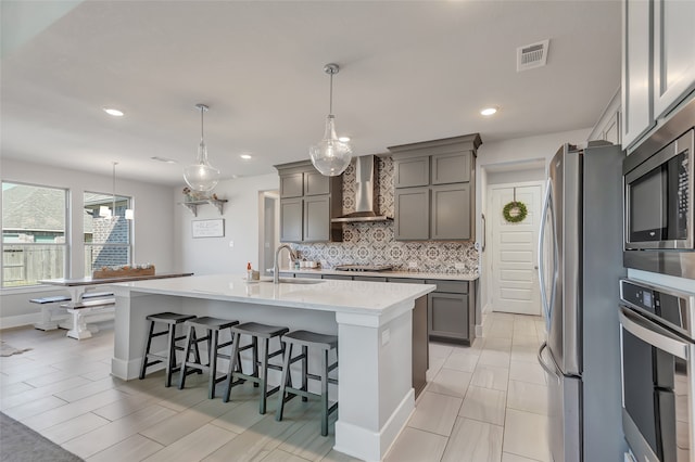 kitchen with appliances with stainless steel finishes, pendant lighting, wall chimney exhaust hood, and an island with sink