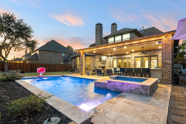 pool at dusk featuring an in ground hot tub, pool water feature, ceiling fan, a bar, and a patio