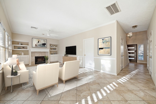 living room featuring a tile fireplace, ceiling fan with notable chandelier, built in shelves, light tile patterned floors, and ornamental molding