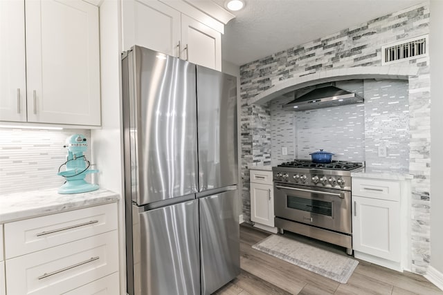 kitchen featuring white cabinetry, light stone countertops, wall chimney range hood, backsplash, and appliances with stainless steel finishes