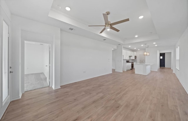 unfurnished living room featuring a tray ceiling, ceiling fan, sink, and light wood-type flooring