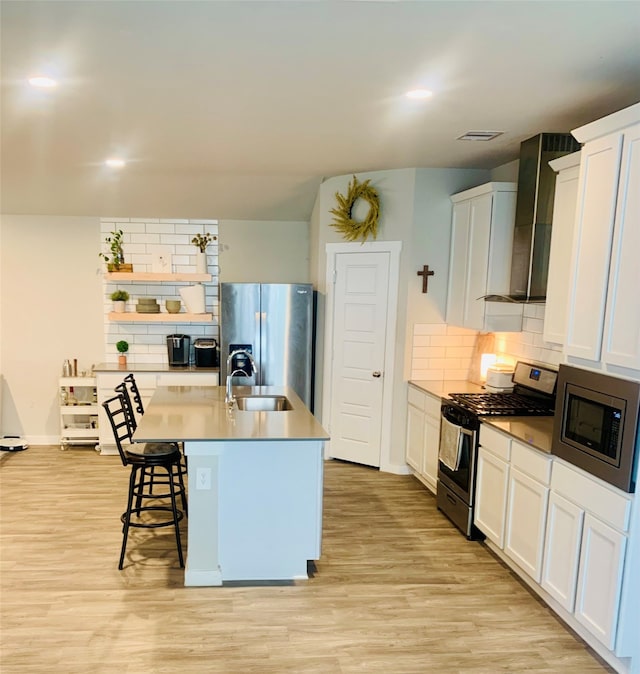 kitchen with wall chimney range hood, appliances with stainless steel finishes, a center island with sink, white cabinets, and light wood-type flooring