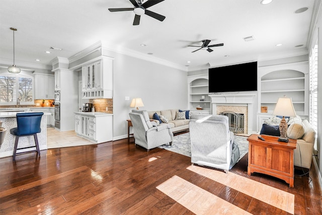 living room featuring ceiling fan, a premium fireplace, built in features, crown molding, and wood-type flooring