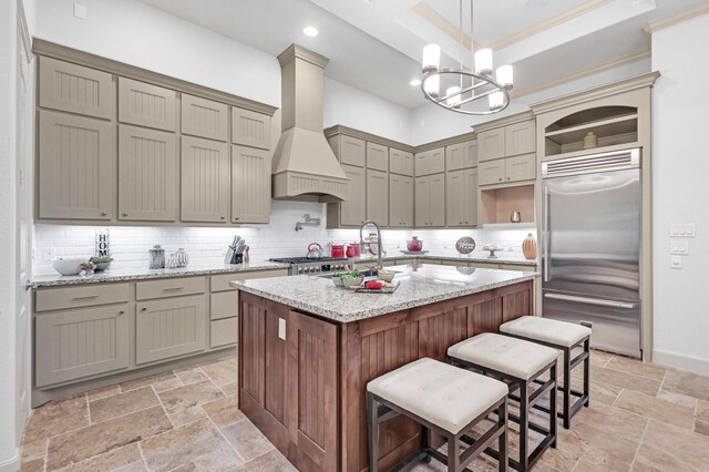 kitchen with a kitchen island with sink, light stone counters, decorative backsplash, stainless steel built in fridge, and custom exhaust hood