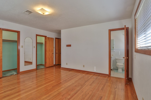 unfurnished bedroom featuring a textured ceiling, light wood-type flooring, two closets, and ensuite bath