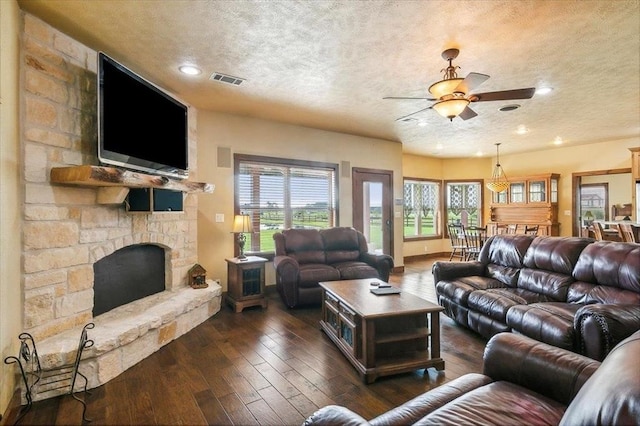 living room with ceiling fan, a stone fireplace, dark hardwood / wood-style flooring, and a textured ceiling