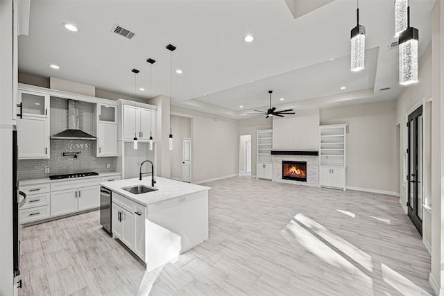 kitchen with wall chimney exhaust hood, decorative light fixtures, a tray ceiling, a kitchen island with sink, and white cabinets