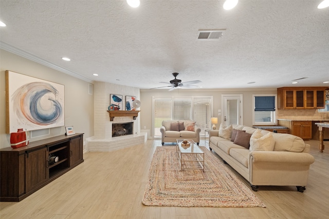 living room with ceiling fan, crown molding, light hardwood / wood-style floors, a textured ceiling, and a fireplace