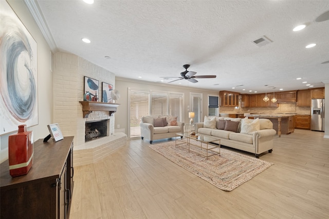 living room featuring ceiling fan, a brick fireplace, light hardwood / wood-style flooring, a textured ceiling, and ornamental molding