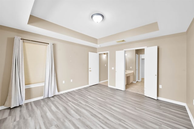 unfurnished bedroom with light hardwood / wood-style flooring, a tray ceiling, and ensuite bath