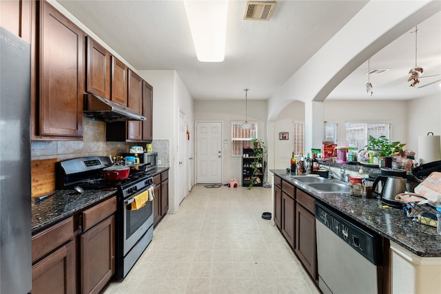 kitchen with appliances with stainless steel finishes, dark stone counters, ceiling fan, sink, and hanging light fixtures