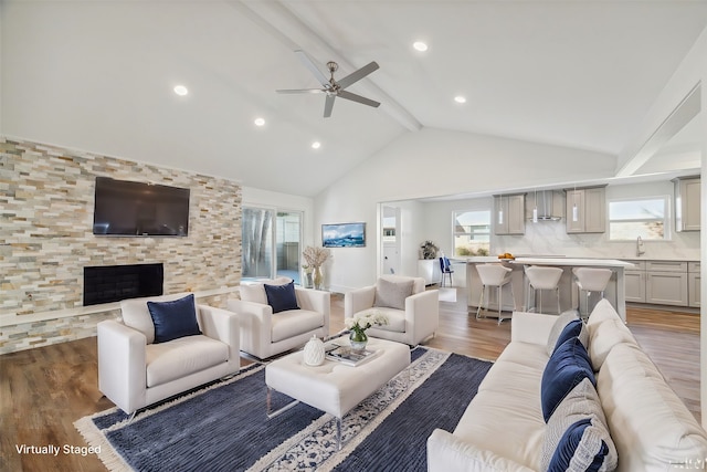 living room with ceiling fan, sink, wood-type flooring, vaulted ceiling with beams, and a stone fireplace