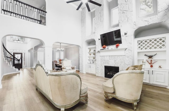 living room featuring built in shelves, a high ceiling, light hardwood / wood-style floors, a fireplace, and ceiling fan with notable chandelier