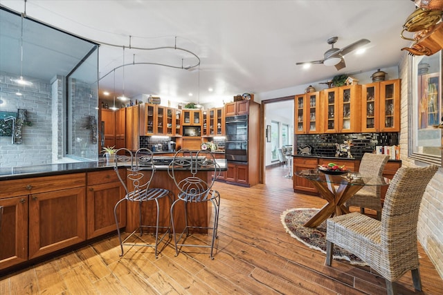 kitchen with black double oven, an island with sink, light wood-type flooring, ceiling fan, and a kitchen breakfast bar