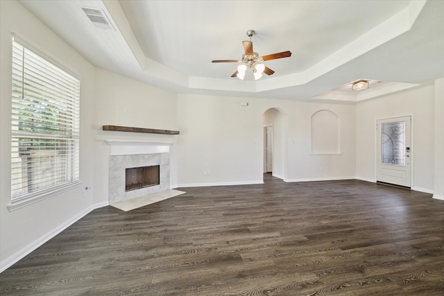 unfurnished living room with a tray ceiling, a tiled fireplace, ceiling fan, and dark wood-type flooring