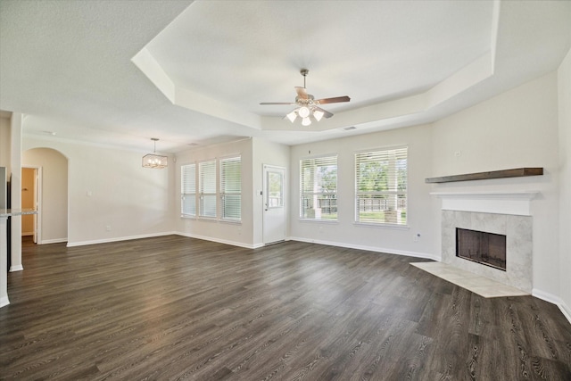 unfurnished living room featuring a tiled fireplace, dark hardwood / wood-style floors, ceiling fan with notable chandelier, and a tray ceiling