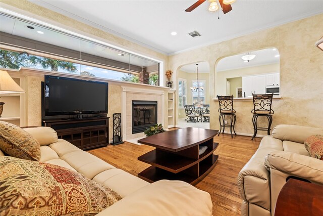 living room with a tiled fireplace, crown molding, light hardwood / wood-style flooring, and ceiling fan with notable chandelier