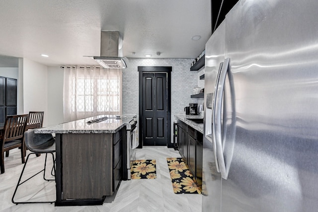 kitchen featuring light stone countertops, wall chimney exhaust hood, brick wall, a kitchen bar, and appliances with stainless steel finishes