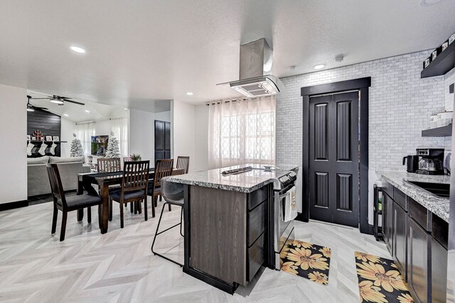 kitchen featuring stainless steel electric range, a kitchen island with sink, exhaust hood, light stone countertops, and dark brown cabinetry