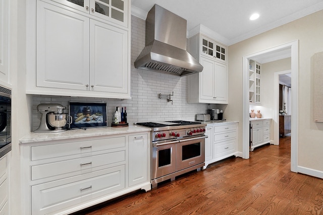 kitchen with wall chimney exhaust hood, dark hardwood / wood-style flooring, range with two ovens, white cabinets, and ornamental molding