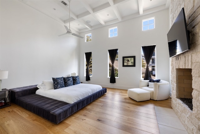 bedroom with beam ceiling, coffered ceiling, a stone fireplace, a towering ceiling, and light hardwood / wood-style floors