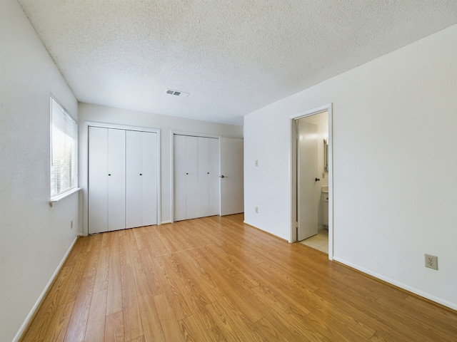 unfurnished bedroom with a textured ceiling, light wood-type flooring, ensuite bathroom, and multiple closets