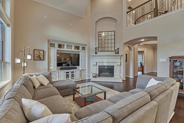 living room featuring arched walkways, a glass covered fireplace, ornamental molding, wood finished floors, and a high ceiling