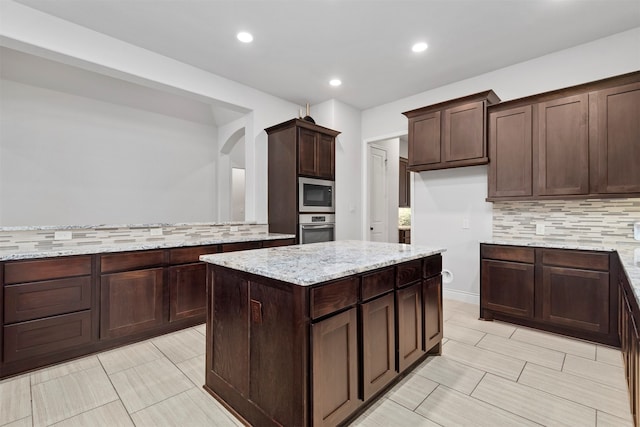 kitchen with dark brown cabinetry, light stone countertops, backsplash, a kitchen island, and appliances with stainless steel finishes