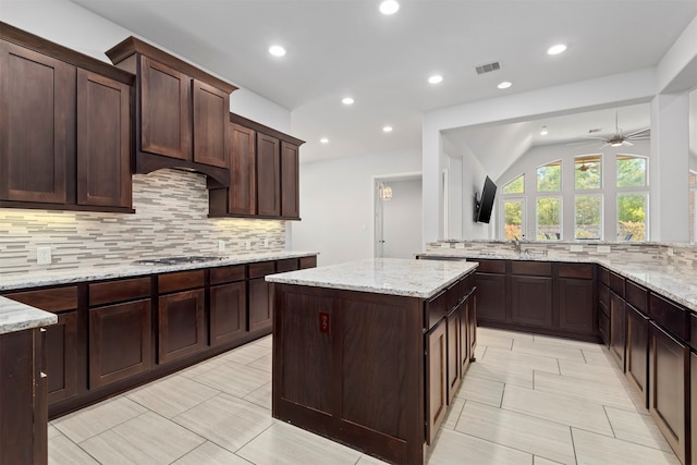 kitchen featuring dark brown cabinetry, ceiling fan, a center island, light stone countertops, and decorative backsplash