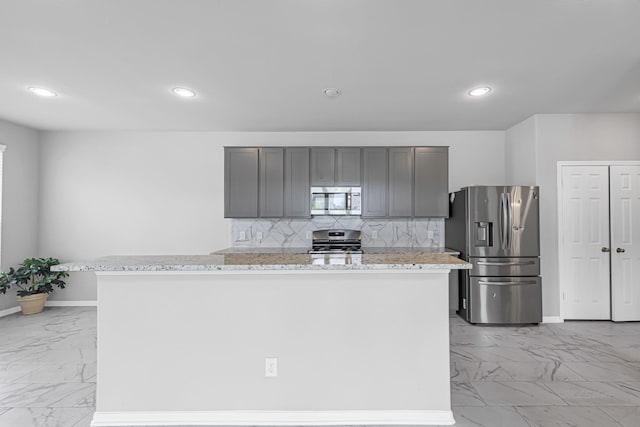 kitchen featuring gray cabinetry, appliances with stainless steel finishes, an island with sink, light stone countertops, and decorative backsplash