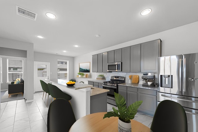 kitchen with decorative backsplash, gray cabinets, light stone countertops, a kitchen island, and stainless steel appliances