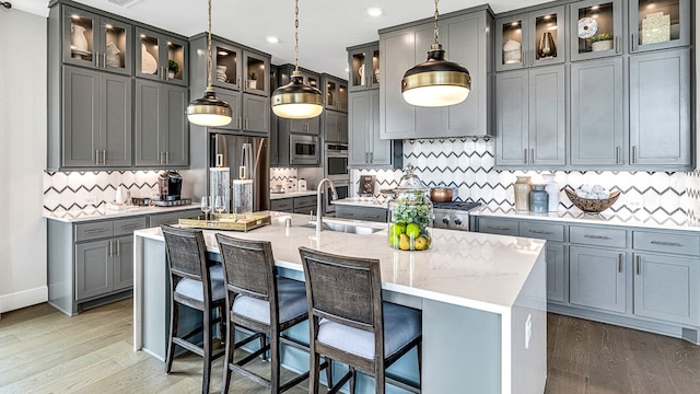 kitchen with tasteful backsplash, stainless steel appliances, a kitchen island with sink, wood-type flooring, and hanging light fixtures