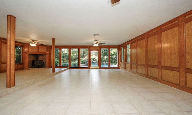 unfurnished living room featuring a textured ceiling, ceiling fan, wooden walls, a wood stove, and light tile patterned flooring