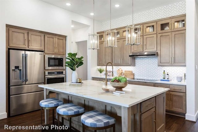 kitchen featuring light stone countertops, decorative backsplash, stainless steel appliances, a kitchen island with sink, and decorative light fixtures