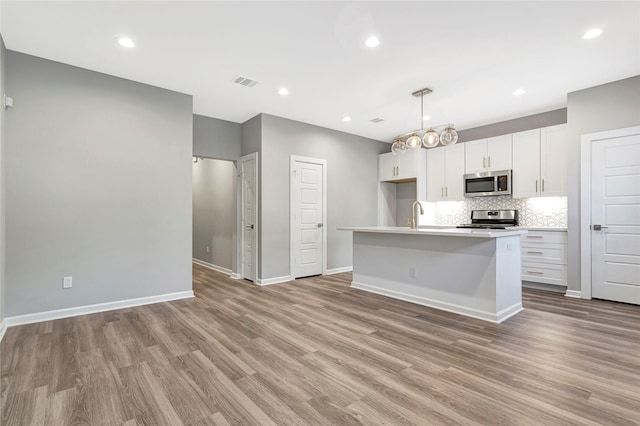 kitchen with white cabinetry, hanging light fixtures, stainless steel appliances, a kitchen island with sink, and light wood-type flooring